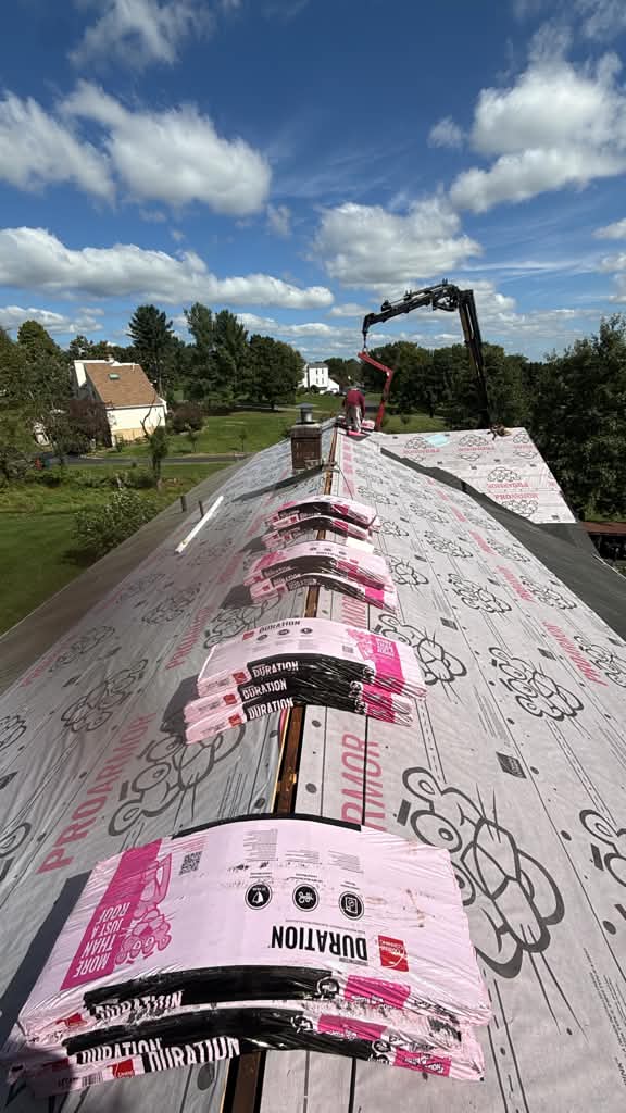 Bundles of new shingles staged on a roof ridge over synthetic underlayment, with a boom truck delivering materials